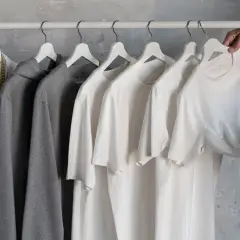 En vit skjorta, grå hoodie och flera vita t-shirts hänger på en klädställning. En hand plockar ner en av t-shirtarna, som en del av en basgarderob herr.