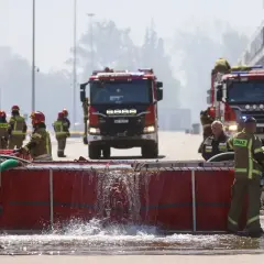 Strażacy gaszą pożar, a ich zarobki są tematem dyskusji. Woda przelewa się przez czerwony zbiornik.