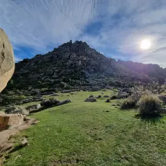 Majestuosa montaña rocosa en la ruta Soto del Real, con un prado verde y el sol brillando en un cielo azul con nubes.