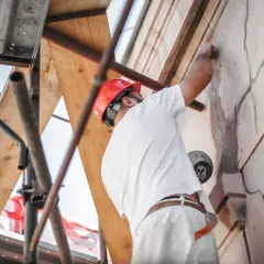 Obrero con casco rojo repara grietas en una fachada desde andamios.