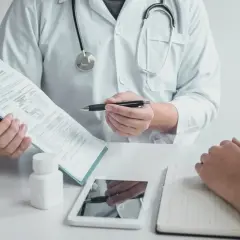 Lekarz w białym kitlu omawia z pacjentem dokumenty, prawdopodobnie dotyczące zwolnienia lekarskiego w Norwegii. Na stole leżą tablet i notatnik.