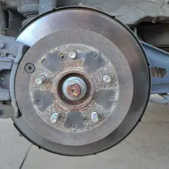 Gros plan sur un disque de frein usé, rouillé et marqué par l'usage. L'étrier de frein est visible, ainsi que des éléments de suspension.