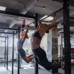 Dwie kobiety wykonują podciąganie na drążku w siłowni. To część treningu, który może być elementem "fran crossfit co to jest".