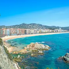 Widok na plażę i zatokę w Lloret de Mar. W oddali widać zabudowania, a na pierwszym planie skaliste wybrzeże i turkusowe morze. Idealne miejsce na wakacje, niedaleko lotniska.