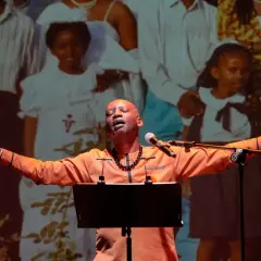A man in an orange shirt sings with his arms outstretched in front of a projection of a family.