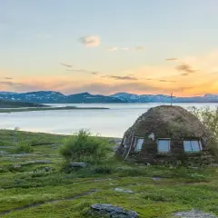 En grästorvstuga med kors på toppen vid en sjö i Padjelanta nationalpark, Sverige, under en solnedgång.