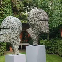 Zwei moderne Gartenskulpturen in Form von Köpfen aus Stein, im Grünen auf Podesten platziert.