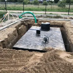 Widok na wykop pod szambo. Duża betonowa płyta, okrągły właz i liczne rury.