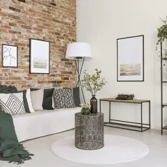Nowoczesne ściany w salonie z cegły, ozdobione grafikami. Wygodna sofa z poduszkami i zielone narzuty tworzą przytulną atmosferę.
