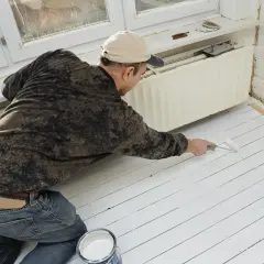 Un homme peint un plancher en bois blanc. Il utilise un rouleau et un pot de peinture. La question "peut on peindre du stratifié" est pertinente pour ce type de projet.