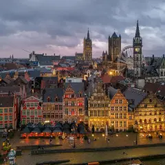 Gryning över Gent, Belgien. Historiska byggnader längs en kanal, med ett pariserhjul och klocktorn som reser sig mot en molnig himmel.