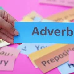 Una mano tiene un biglietto blu con la parola "Adverb". Sullo sfondo, altri biglietti colorati con parole come "Grammar" e "Vocabulary".