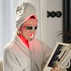 Relaks w domu z maską do terapii światłem LED. Kobieta w szlafroku i ręczniku na głowie czyta książkę, podczas gdy maska emituje czerwone światło.