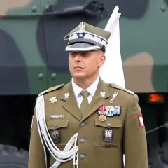 Oficer w mundurze, z orderami na piersi, stoi obok polskiej flagi. Nad nim widać fragment pojazdu wojskowego.
