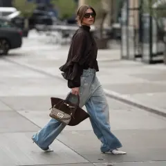 Kobieta w brązowej bluzce z falbanami i jasnych, szerokich spodniach z przetarciami, z brązową torebką. Czy szerokie spodnie są modne? Tak, ten look to dowód!
