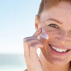 Frau mit Sommersprossen trägt Sonnenfluid auf ihr Gesicht auf, um ihre Haut vor der Sonne zu schützen.