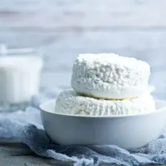 Due forme di ricotta fresca in una ciotola bianca, con un bicchiere di latte sullo sfondo. La ricotta si può congelare per conservarla più a lungo.