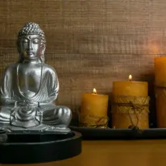 Srebrna figurka Buddy siedząca w medytacji, obok płonące świece na drewnianym tle.