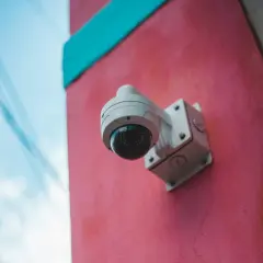 Caméra de surveillance blanche fixée sur un mur rose vif, sous un ciel bleu. Un rappel visuel de la loi sur la caméra de surveillance.
