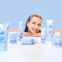 Uśmiechnięta dziewczyna z niebieskimi pasemkami otoczona kosmetykami Face Boom. Wśród nich kremy i sera, które dzięki caprylyl glycol odświeżają skórę.
