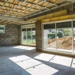 Wnętrze w budowie z betonową podłogą i ścianami. Widoczna konstrukcja sufitu i okna z widokiem na plac budowy. Powstaje nowa ściana działowa.