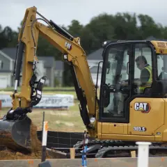 Żółta minikoparka CAT 303.5 pracuje na budowie nowych domów. Jej szerokość pozwala na precyzyjne manewrowanie w ciasnych przestrzeniach.