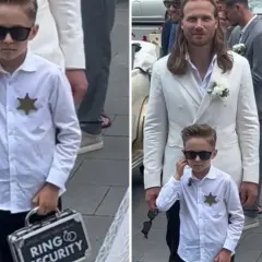 Chłopiec w okularach przeciwsłonecznych, z odznaką i walizeczką "Ring Security", towarzyszy parze młodej.