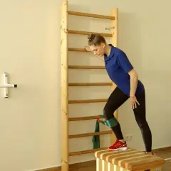 Frau trainiert mit Theraband am Knie an einer Sprossenwand, um die Beinmuskulatur zu stärken.
