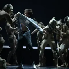 Tancerz w Teatrze Wielkim w Warszawie, otoczony przez postaci w maskach, w scenie z repertuaru.