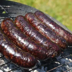 Pięć ciemnych, błyszczących kiełbasek kaszanka na grillu, dymiących na ruszcie nad żarem.