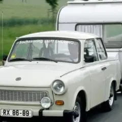 Biały trabant ciągnie przyczepę kempingową po drodze.