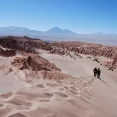 Dos excursionistas caminan por una duna en el desierto de Atacama, con formaciones rocosas y montañas al fondo bajo un cielo azul.