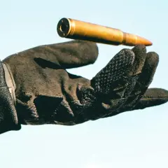 Una mano con guantes tácticos sostiene un cartucho dorado contra un cielo azul claro.