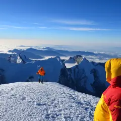 Widok z góry, gdzie są Himalaje. Dwóch alpinistów w kolorowych kurtkach na ośnieżonym szczycie, pod nimi morze chmur i kolejne pasma górskie.