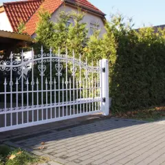 Biała, ozdobna brama wjazdowa, niczym wiejski płot, otwiera drogę do domu z czerwonym dachem, otoczonego zielenią.
