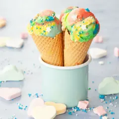 Zwei bunte Eiswaffeln mit Regenbogen-Eis und Streuseln stehen in einem hellblauen Becher. Marshmallows in Herz- und Wolkenform liegen verstreut.