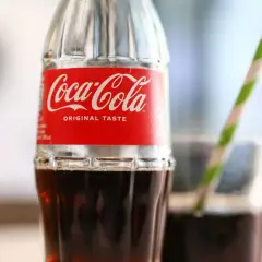 Szklana butelka Coca-Coli z czerwono-białą etykietą i słomką w tle.