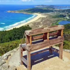 Banco de madera en un monte ventoso con vistas a una playa, el mar azul y un lago.