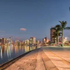 Wieczorny widok na nowoczesną panoramę miasta, gdzie odbijają się światła. To Luanda, stolica Angoli, z palmami na pierwszym planie.