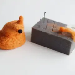 Pomarańczowy stworek z filcu na sucho, obok gąbka z igłami i kłębki wełny.