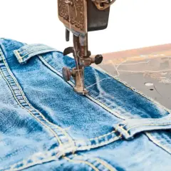 A sewing machine stitches a classic jean stitch onto blue denim fabric, highlighting the durable construction of jeans.