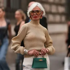 Elegancka kobieta po 60-tce w beżowym golfie i okularach, z zieloną torebką. Inspiracja dla mody dla 60 latki.