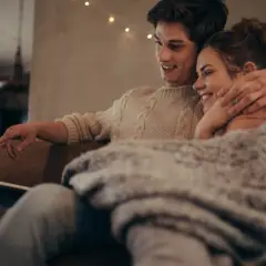 Para młodych ogląda coś na laptopie, przytuleni pod kocem. Ich bliskość i uśmiechy sugerują, że historia o "fake dating" tak bardzo angażuje emocjonalnie czytelników, bo pozwala przeżywać rodzące się uczucia.