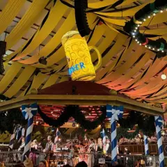 Wielki, świecący kufel piwa wisi nad sceną z zespołem. Dekoracje w kolorach biało-niebieskim i żółtym tworzą atmosferę Oktoberfest 2025.