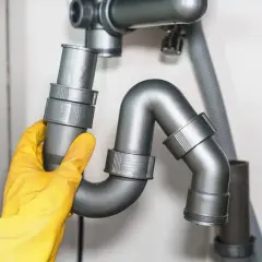 Main d'un plombier en gant jaune ajustant un siphon de rejet d'eau PVC sous un évier.