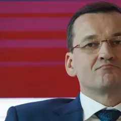 Mateusz Morawiecki w garniturze i okularach na tle czerwono-fioletowych pasów.