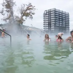 Ludzie relaksują się w parujących basenach termalnych w Karlowych Warach. W tle nowoczesny budynek i drzewa.
