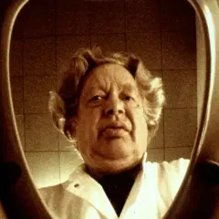 En man med lockigt hår, klädd i vit rock, tittar in i en toalettstol. Ernst Hugo Järegård i en minnesvärd scen.