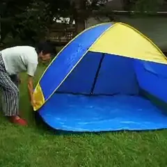 Osoba pochyla się nad rozkładanym namiotem plażowym w kolorze niebiesko-żółtym, stojącym na trawie.
