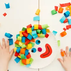 Bambini creano lavoretti di carnevale da appendere: un pagliaccio con capelli colorati su un piatto di carta, circondato da coriandoli di carta.